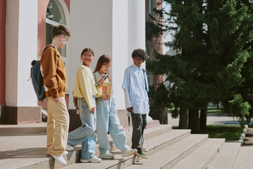 Group of multiethnic teenagers standing on steps outside educational building, smiling and talking together, one girl holding smartphone while others engaging in conversation