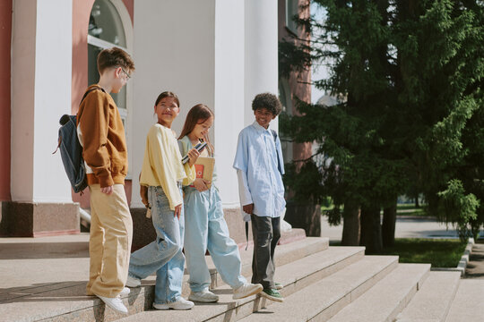 Group of multiethnic teenagers standing on steps outside educational building, smiling and talking together, one girl holding smartphone while others engaging in conversation
