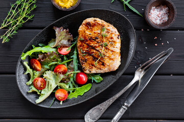 Grilled pork chop with fresh salad in black plate on black wooden background, meat top view
