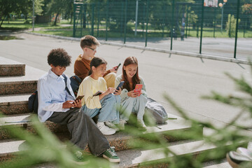 Group of multiethnic teenagers sitting on outdoor steps using smartphones and tablet, Caucasian boy, Black boy, Asian girl, Caucasian girl engaging with digital devices together