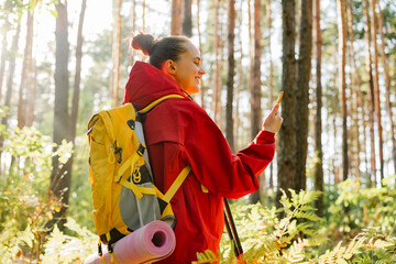 A smiling female hiker in a red jacket with a yellow backpack stops to check her smartphone while trekking through a peaceful, sun-drenched forest.
