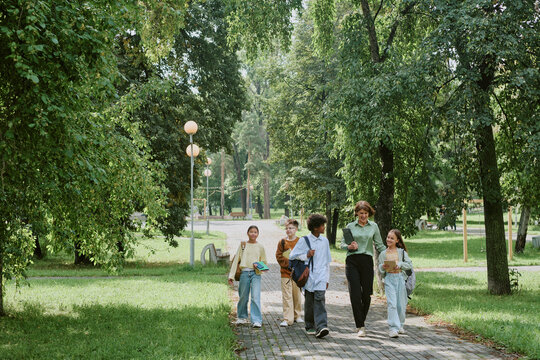 Group of diverse teenagers walking together outdoors in park, carrying backpacks and books, smiling and talking, surrounded by green trees and grass, enjoying time after school