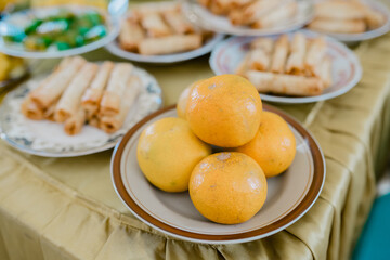 Fresh oranges neatly stacked on a ceramic plate, placed on a dining table. Traditional food arrangement with fruits and snacks in the background, often served during gatherings, celebrations.