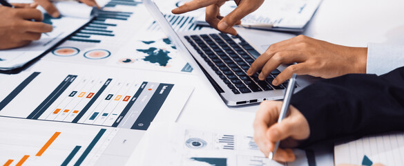 A diverse business team collaborates on a digital marketing strategy. Laptops and documents filled with data, charts, and graphs fill the modern office space. SACTR