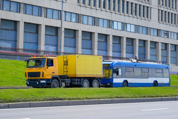 An orange flatbed tow truck hauls a damaged blue and white trolley bus on the side of a road in front of a building. The trolley bus appears to be disabled on the grass