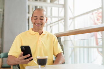 Mature Hispanic bald man sitting at a cafe while using phone