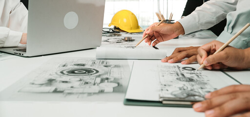 A modern office scene showing engineers collaborating over blueprints and designs while utilizing laptops. Hard hat and sketches highlight project planning and teamwork. SACTR © InfiniteFlow