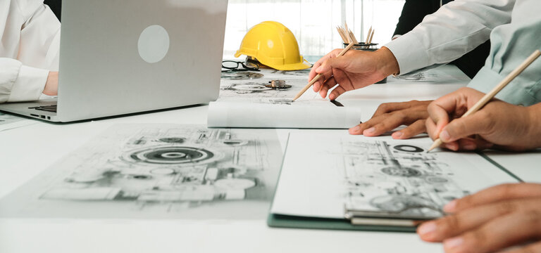 A modern office scene showing engineers collaborating over blueprints and designs while utilizing laptops. Hard hat and sketches highlight project planning and teamwork. SACTR
