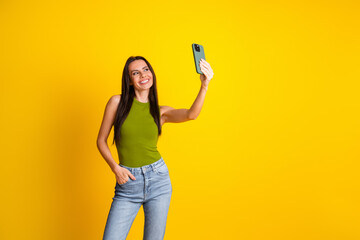 Happy woman in green top holding smartphone against vibrant yellow background, making selfie, showing cheerful expressions