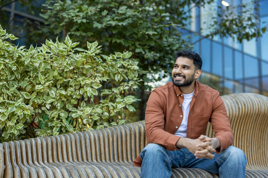 Smiling young man seated outdoors surrounded by greenery and modern architecture
