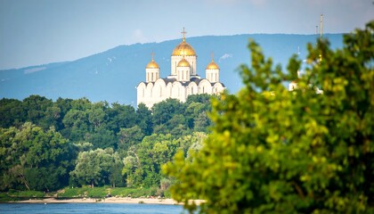 Panoramic view of a majestic white cathedral nestled amidst lush greenery, with golden domes gleaming under a clear sky.