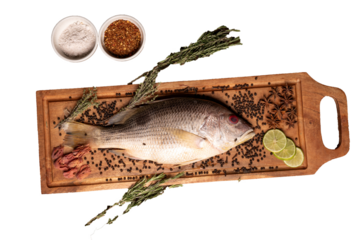 Top view of raw red snapper fish with whole spices, herbs, and lemon slices on wooden tray, styled with chili flakes and salt, isolated PNG for culinary presentation.