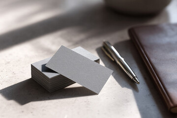Clean, professional layout with a stack of grey business cards, a sleek pen, and notebook on a textured surface under sunlight. Perfect for branding, design, and office themes.