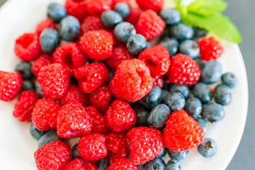 Detailed macro view of ripe raspberries and blueberries with mint on a white plate. Bright natural colors highlight organic fruit rich in antioxidants and vitamins.