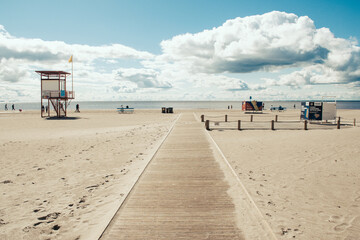Wide, sandy beach with a wooden walkway leading to the shore. There are lifeguard stations and people walking along the beach. The sky is partly cloudy with scattered clouds