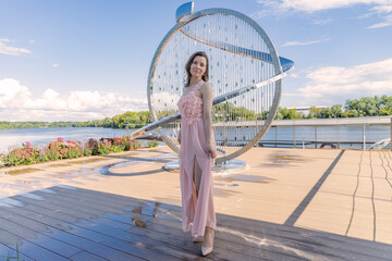 Full body portrait of a young woman in a flowing pink dress standing gracefully in front of a shiny circular modern sculpture on a riverside boardwalk