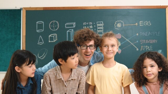 Caucasian professional teacher sitting while hugging diverse cute student at class and looking at camera. Group of smart multicultural children smiling to camera while standing at classroom. Pedagogy.