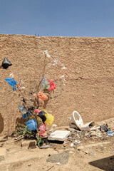 Dry bush entangled with colorful plastic bags near a rough beige wall, surrounded by urban litter and broken ceramic toilet pieces under a clear blue sky in a deserted area.