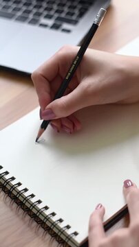 Tight ecu overhead video of hands writing with a graphite pencil in a white spiral notebook on a clean wooden desk, with a silver laptop in blurred bokeh background. Focus, clarity, and productivity