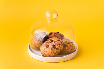 Stack of fresh chocolate American style chip cookies with nuts and chocolate drops on a tray with transparent lid on yellow background. Cookies with peanut butter filling inside a glass jar