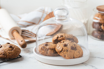 Stack of fresh chocolate American style chip cookies with nuts and chocolate drops on a tray with transparent lid on white marble background. Cookies inside a glass jar with kitchen utensils