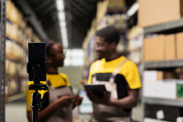African american staff members filming warehouse operations for e-commerce activity, industrial racks in depot. Colleagues vlogging ad for large scale online shipping tasks, promoting delivery.