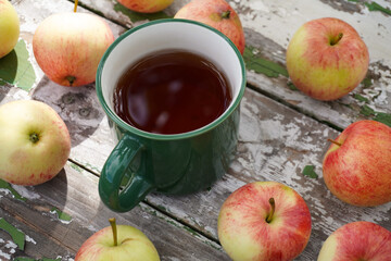 Green enamel mug with hot tea surrounded by ripe red and yellow apples on rustic wooden table