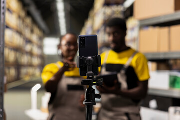 Black team of colleagues recording a promotional vlog in a storehouse, presenting goods in pallets on big industrial racks. Working on a commercial to demonstrate order fulfillment.