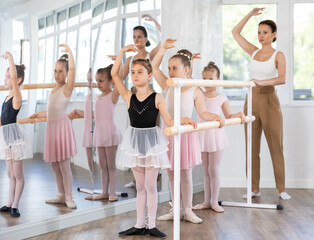 Obraz premium Adult female teacher corrects group of girls dancers standing in first position at ballet barre