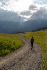 Obraz premium Back view of a solitary hiker on a trail, with a stormy sky but light illuminating the landscape. Represents solitude and nature.