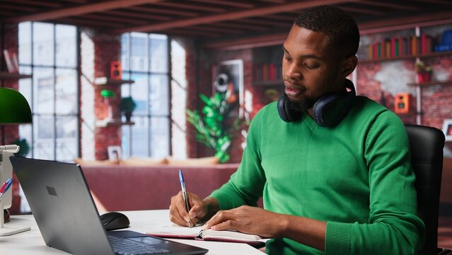 Smiling black man taking notes while watching a tutorial presentation, doing online self learning and writing details. Young adult freelancer writes in his textbook notepad. Camera B.
