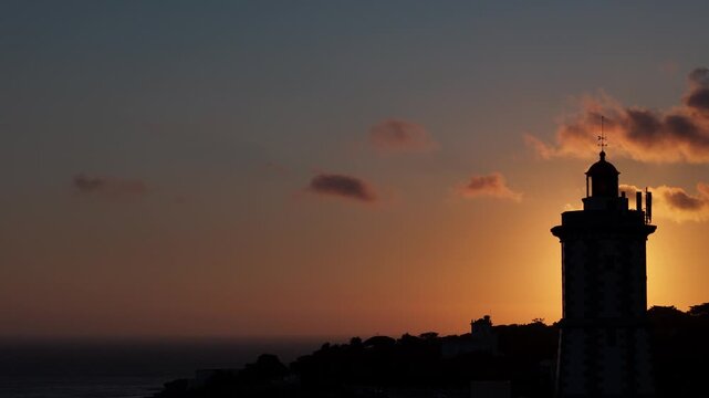 Cinematic backlit view of sunset view behind a silhouette of a lighthouse on a coastal cliff in Cascais, Portugal with dramatic moving clouds