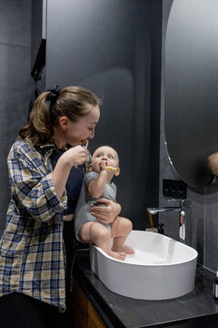 A woman with her baby brushes her teeth while holding a baby in hands in the bathroom