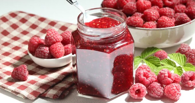 Woman taking delicious raspberry jam from glass jar with spoon at white table, closeup