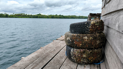 Stack of worn tires covered with barnacles on a wooden pier by calm water under a cloudy sky.