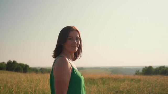 confident woman in green gown stands tall in sunlit grassy field gazing ahead with calm focused expression under warm sky, serene rural landscape stretches behind as golden grass sways gently