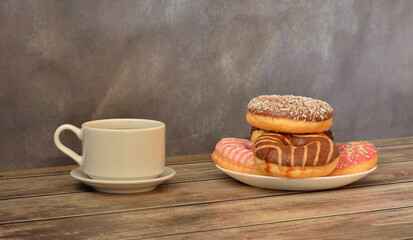 A cup of hot coffee and a plate with several fresh donuts in multi-colored glaze on a wooden table.