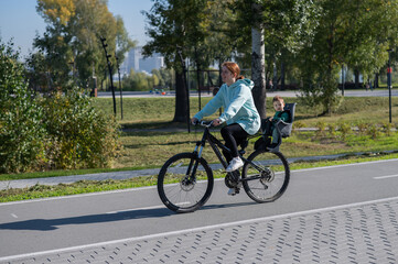 Obraz premium Caucasian woman riding bicycle with toddler son sitting behind her in child seat in park. 