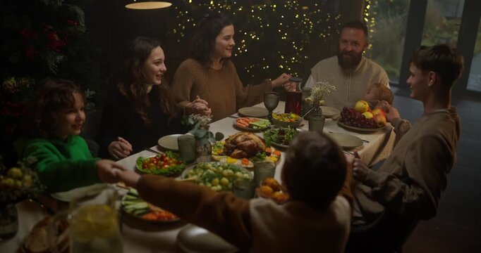 Happy family husband wife and their children sitting at the table holding hands and praying before eating on Christmas Day at the table