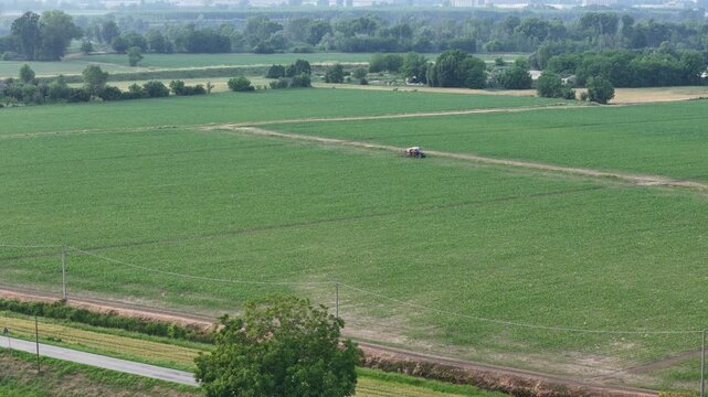 erial shot of a tractor with boom sprayer moving through a vast maize monoculture, applying chemical treatments associated with GMO crop production and environmental sustainability concerns.