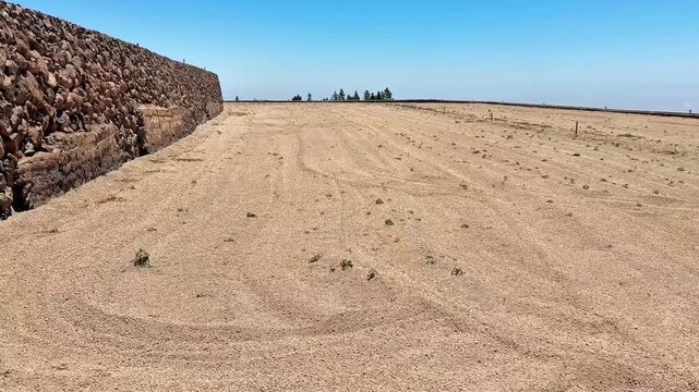 A vast dry expanse showcasing a robust stone wall and limited vegetation beneath a clear, bright blue sky