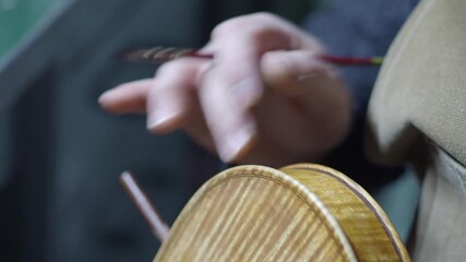 Skilled violinmaker applies antiquing to the marbled ribs and purfling of a violin, revealing the flamed maple pattern on the back plate in workshop setting, captured in close-up detail shot.