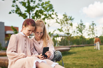 Two Caucasian girls sitting closely on bench using smartphone together outdoors, one wearing glasses, showing focused expression