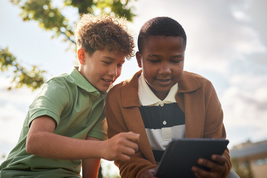 Two boys, one Caucasian and one Black, both children, sitting outdoors interacting with digital tablet, smiling and focusing on screen - Powered by Adobe