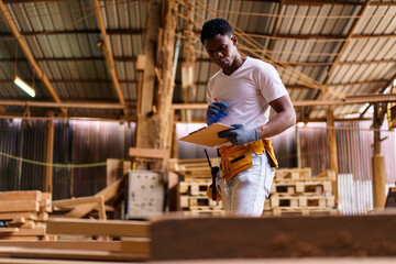 Obraz premium Black carpenter working in woodworking factory, carefully inspecting a wooden plank after cutting. Concept of craftsmanship, labor, and skilled profession.