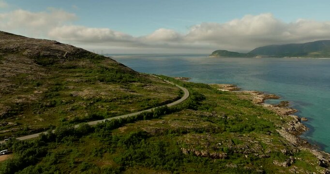 Aerial view of cars tracing a curving coastal road on Kvaloya island in Troms, with fjord water and rocky shoreline under soft clouds on a bright summer day.