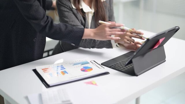Two people are sitting at a desk with a laptop and a stack of papers. One of them is pointing at the laptop screen