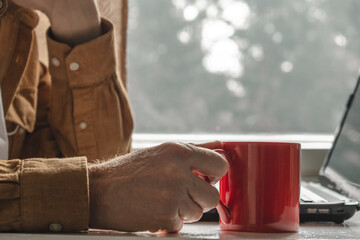 person casually grips red mug, sitting by window with natural light illuminating room. laptop rests nearby, suggesting moment relaxation during work. close up.