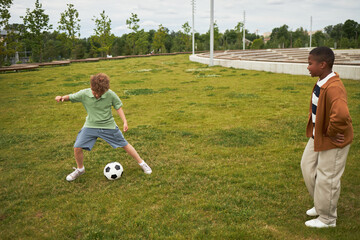 Fototapeta premium Caucasian boy dribbling soccer ball on grass while Black boy watching nearby, both children engaging in outdoor sports activity in park setting during daytime