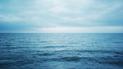 Obraz premium A wide, expansive view of a dark blue ocean meeting a cloudy, pale blue sky. Gentle waves ripple across the water's surface, reflecting the muted light.
